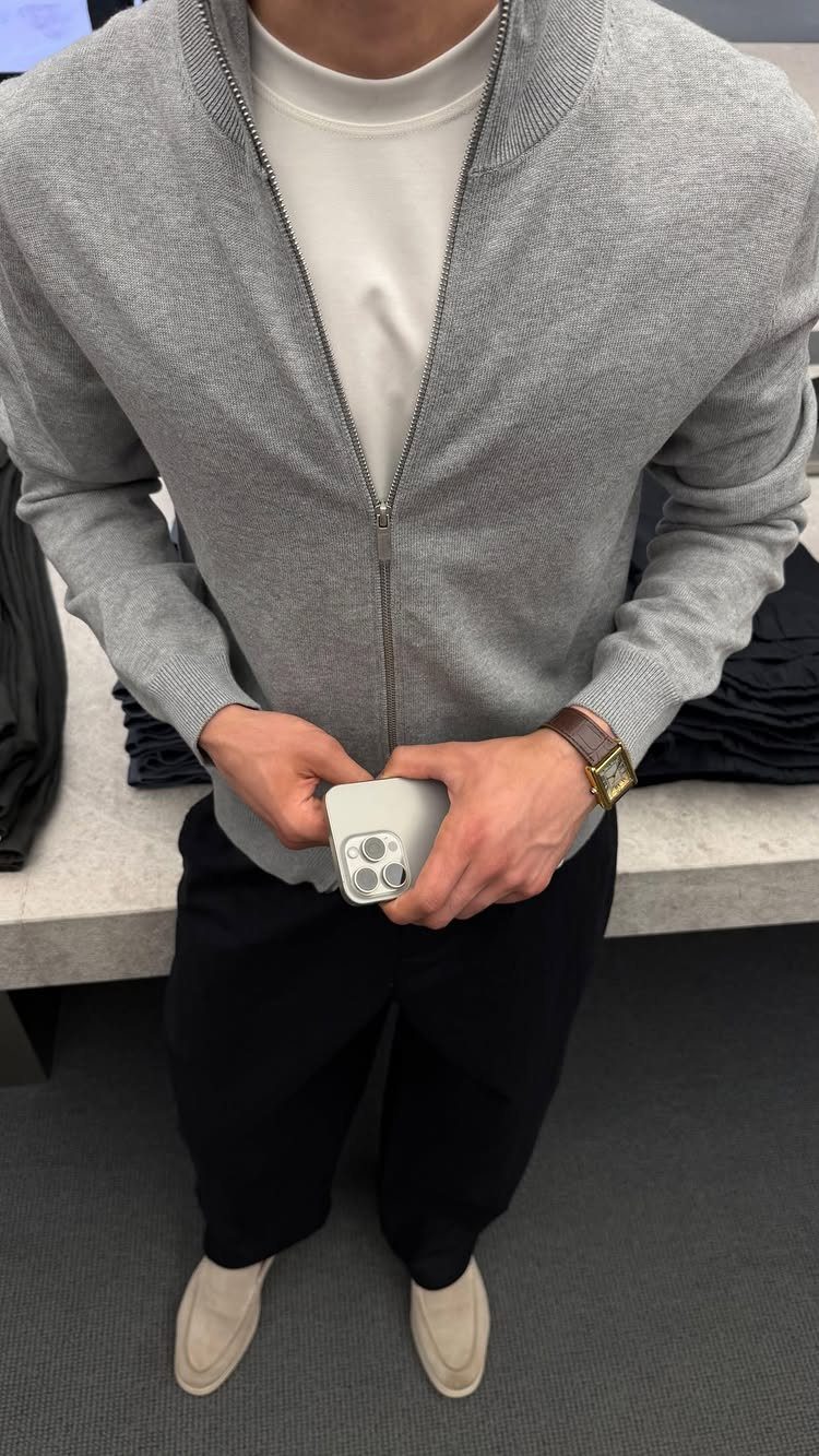 Man wearing a grey zip-up sweater, black trousers, and beige loafers while holding a phone.