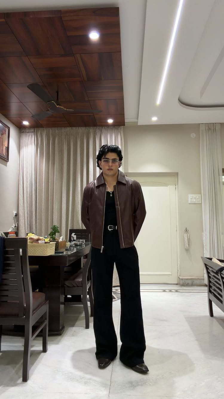 Young man posing in a brown leather jacket and wide-leg black trousers indoors.