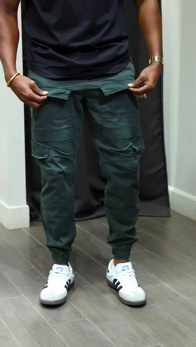 Man wearing dark green cargo joggers and white sneakers standing against a dark background.