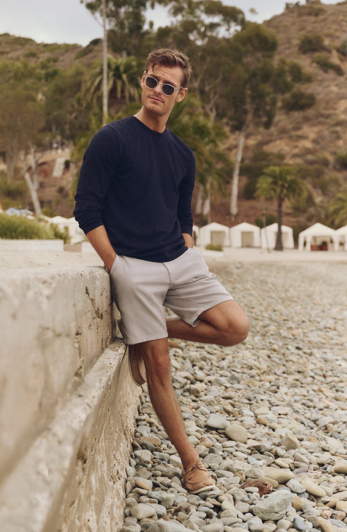 Man in navy shirt and grey chino shorts posing on a rocky beach.