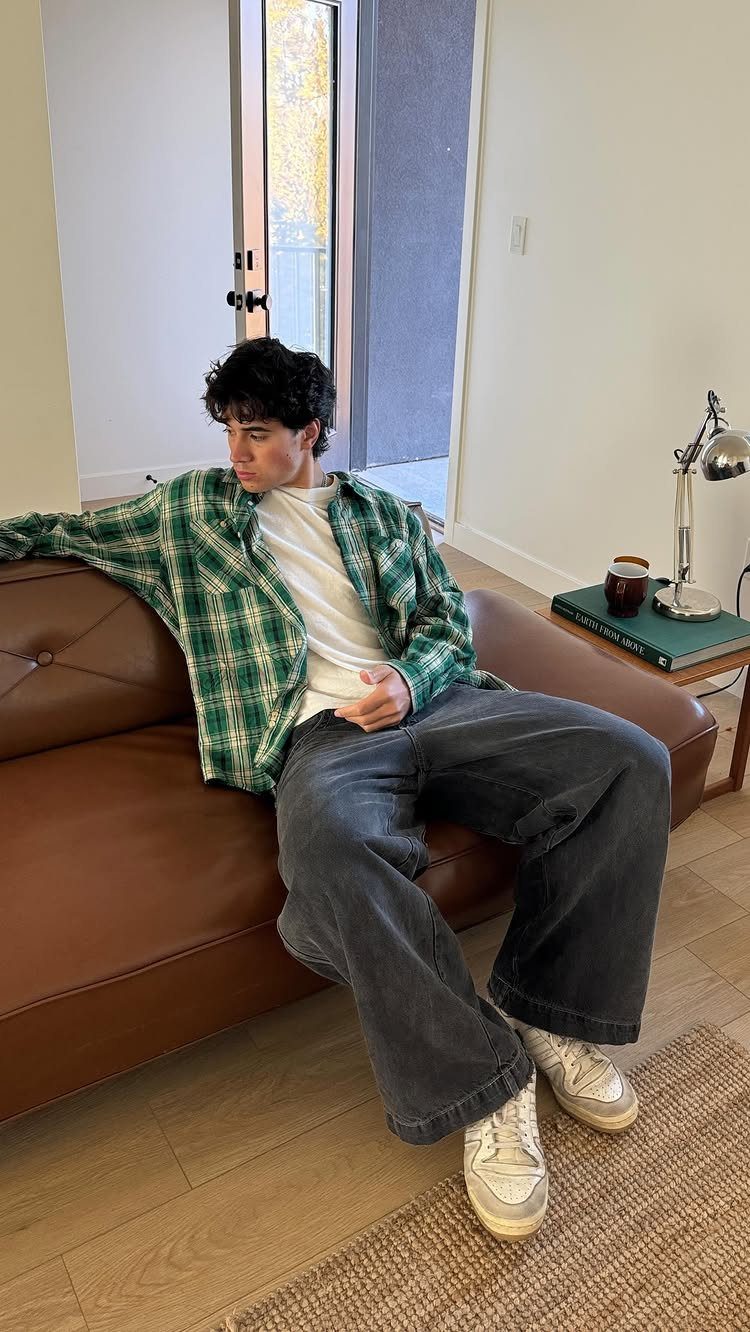 Man sitting on a leather sofa wearing a green plaid flannel shirt and baggy jeans.