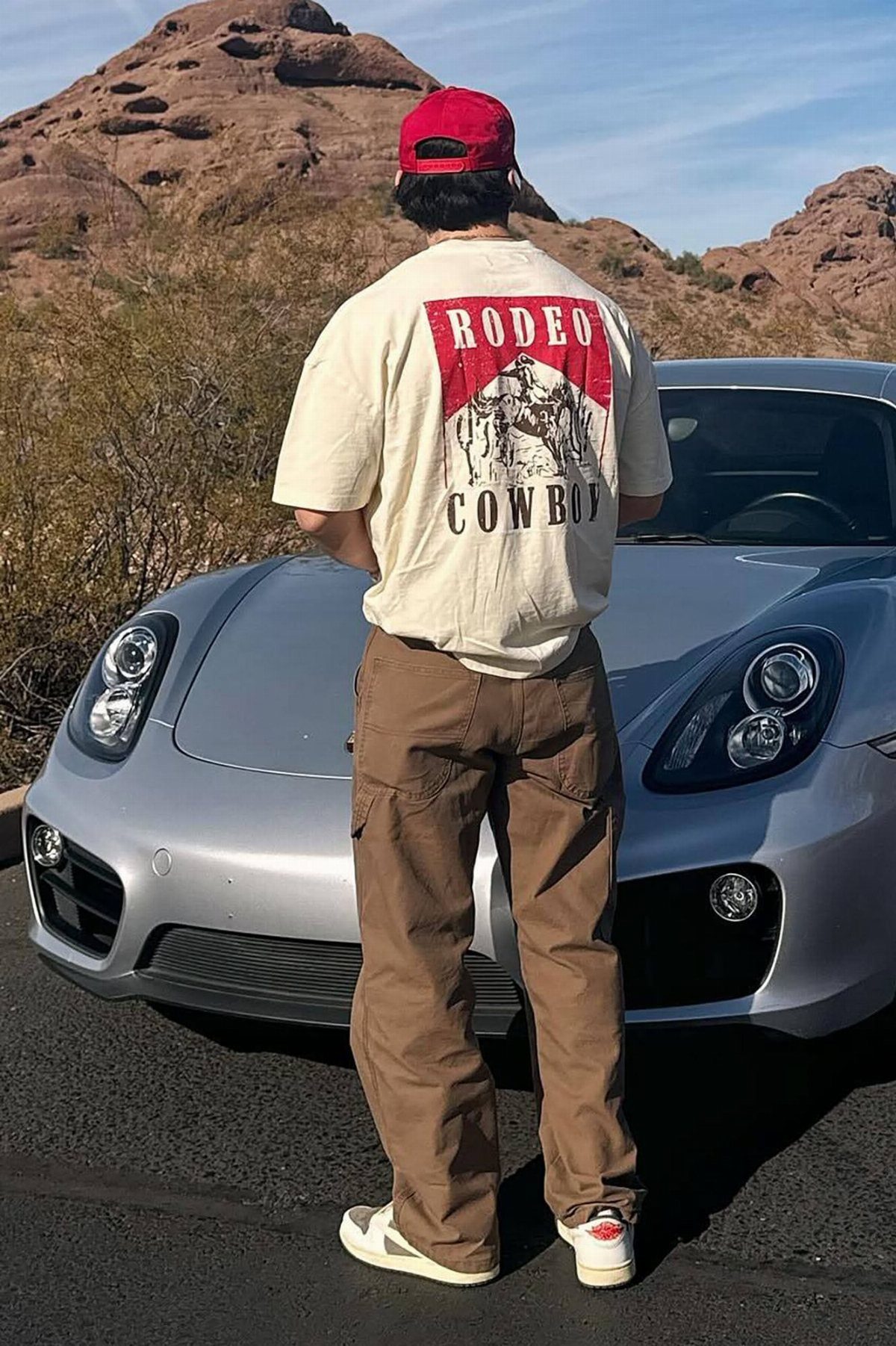 Man wearing a cream rodeo graphic t-shirt and brown cargo pants standing by a silver car.