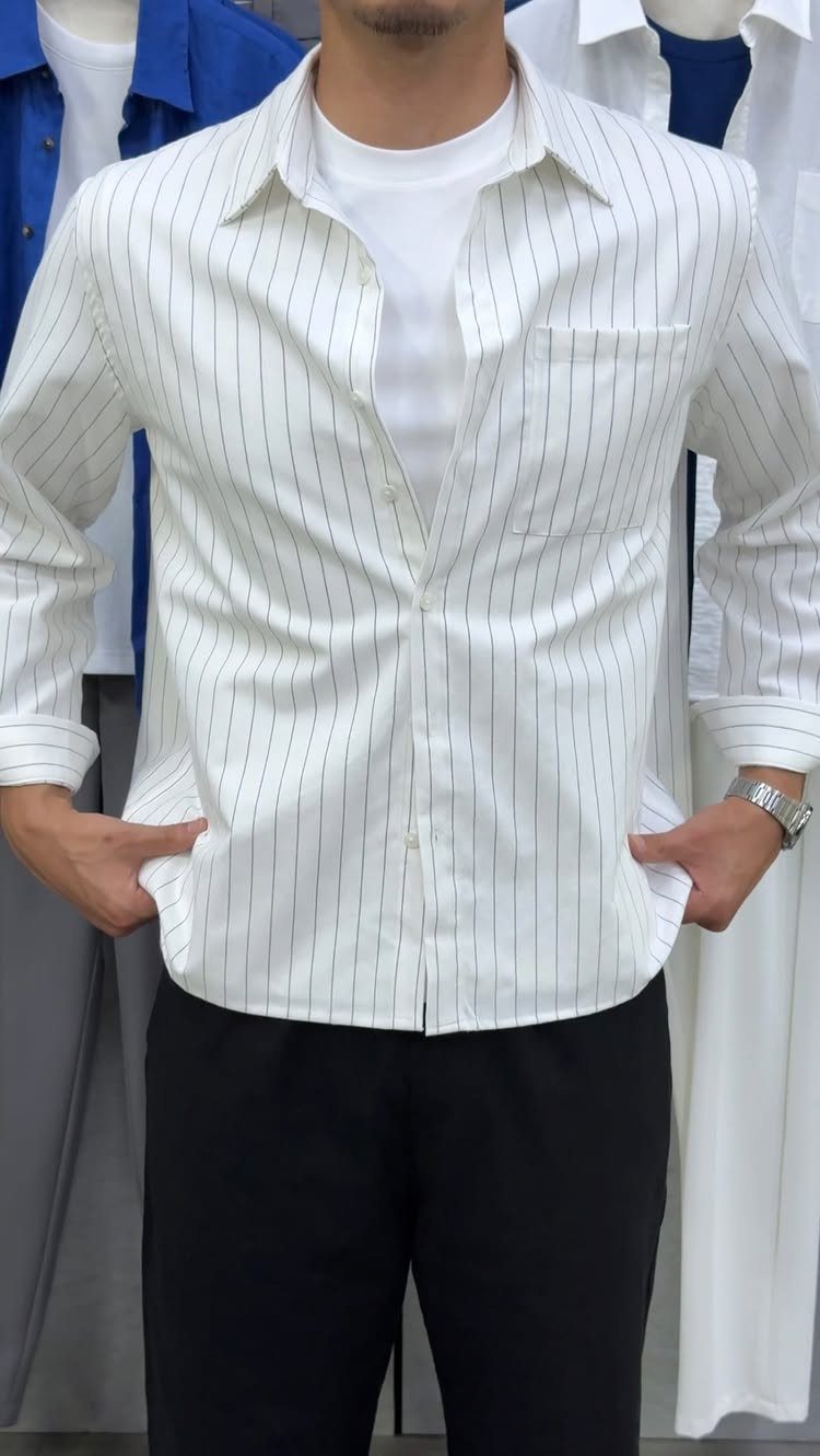 Man wearing a white striped button-down shirt over a white t-shirt and black trousers.