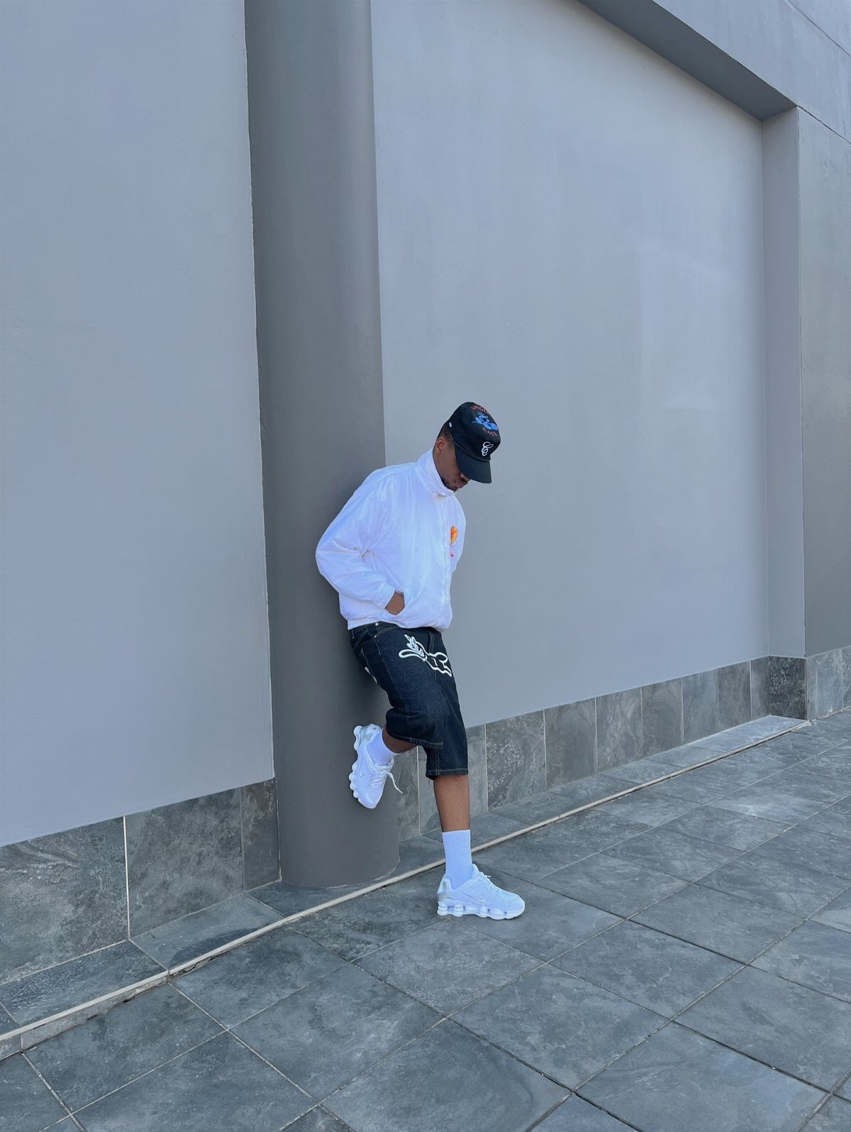 A man in white shirt and black shorts wearing chunky white sneakers against a grey wall.