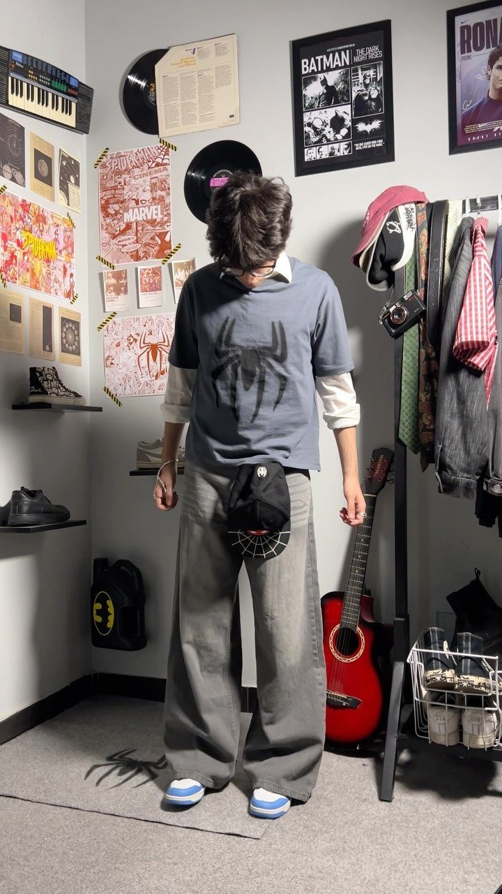 A young man wearing a graphic t-shirt, long sleeves, and wide-leg trousers standing in a room.