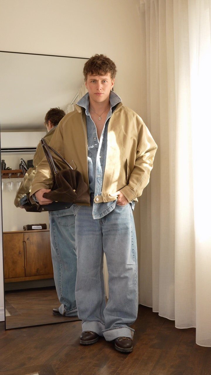 A young man posing in a tan jacket, denim shirt, and wide-leg jeans.
