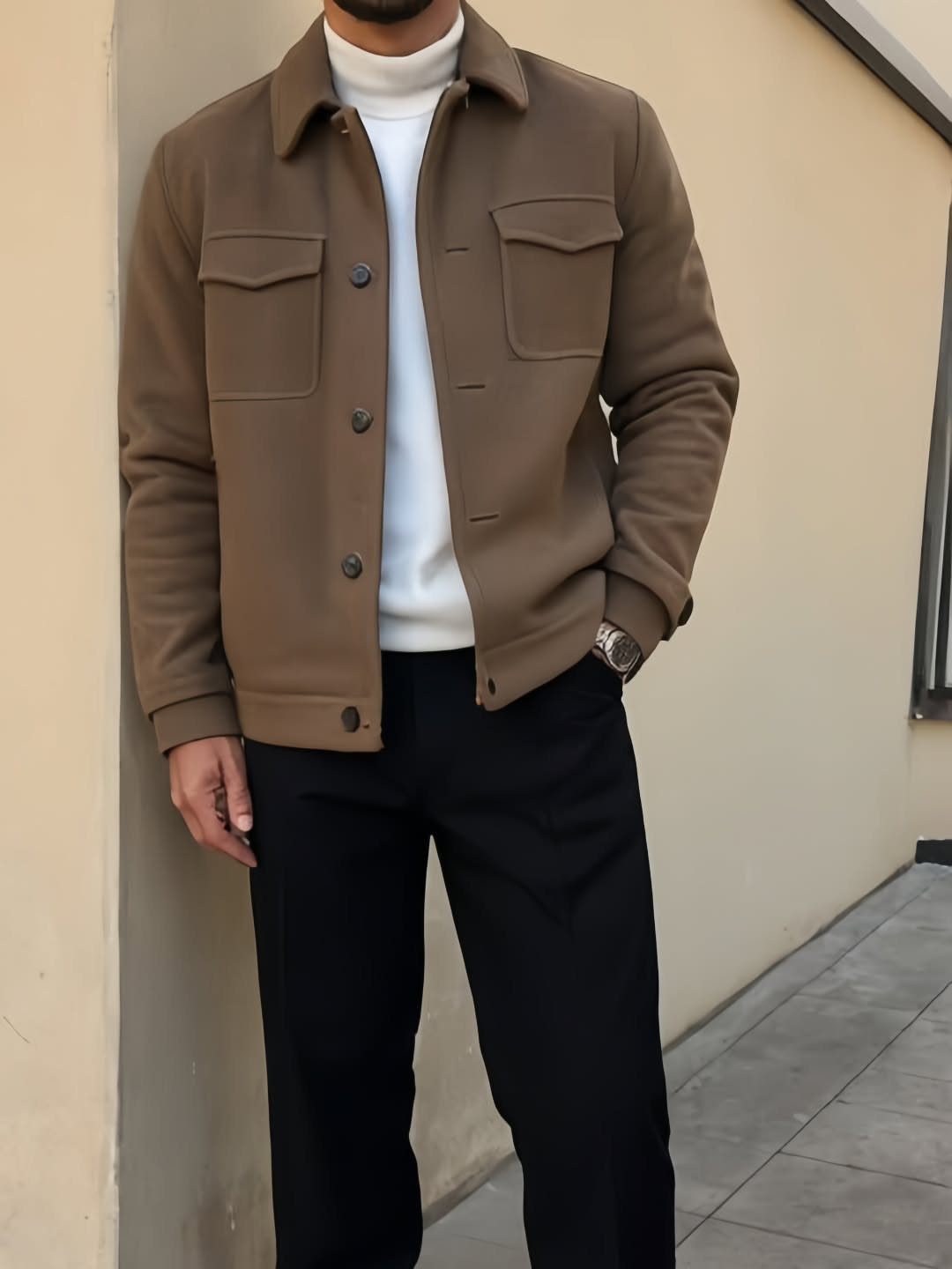 Man wearing a brown collared jacket, white turtleneck, and black trousers standing against a wall.