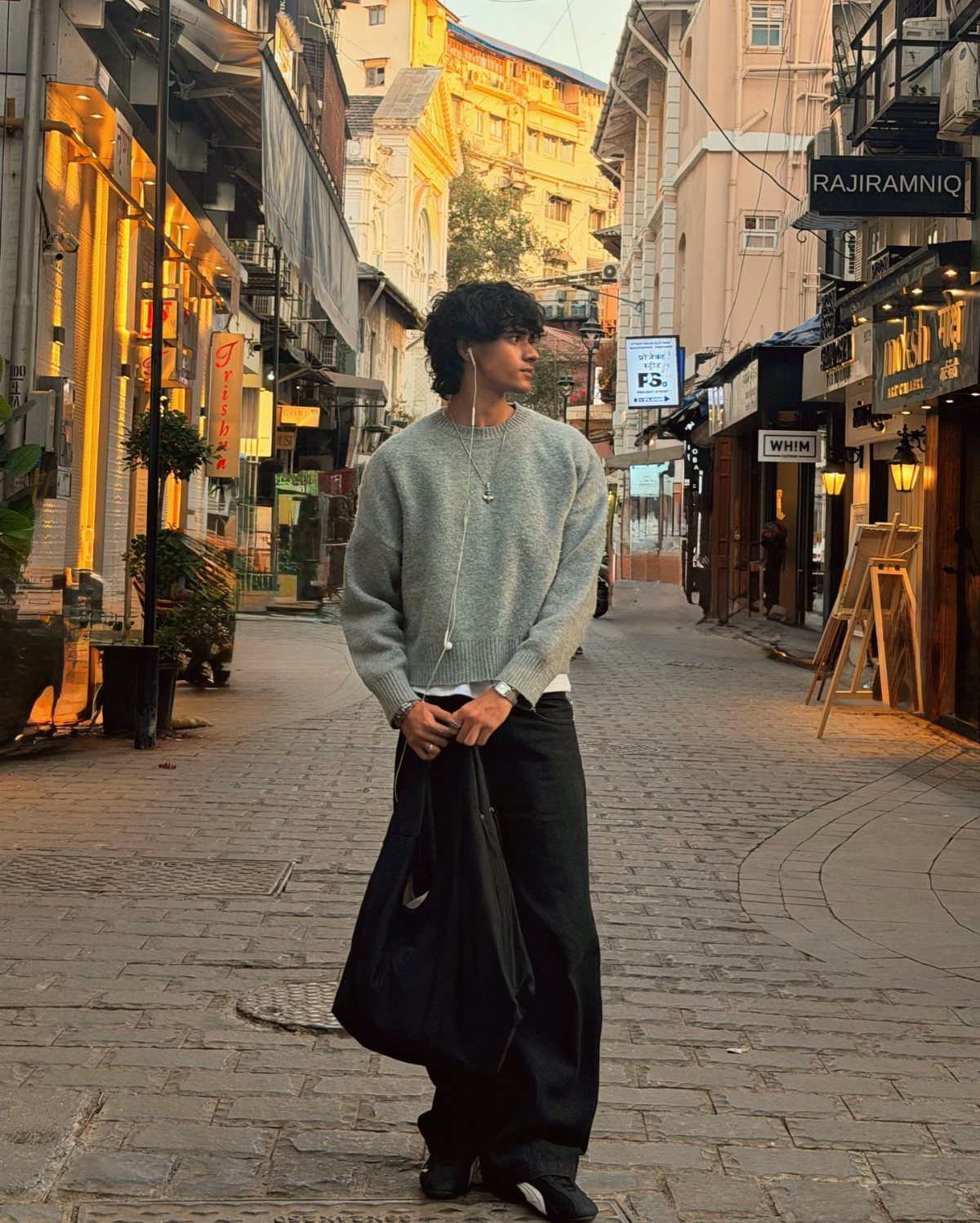 A young man in a grey sweater and black trousers standing on a city street.