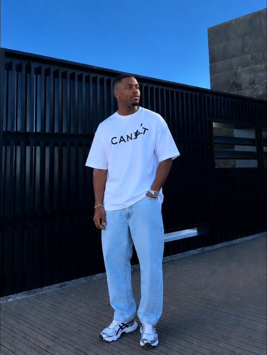 Man in a white graphic t-shirt and light blue jeans standing against a black fence.