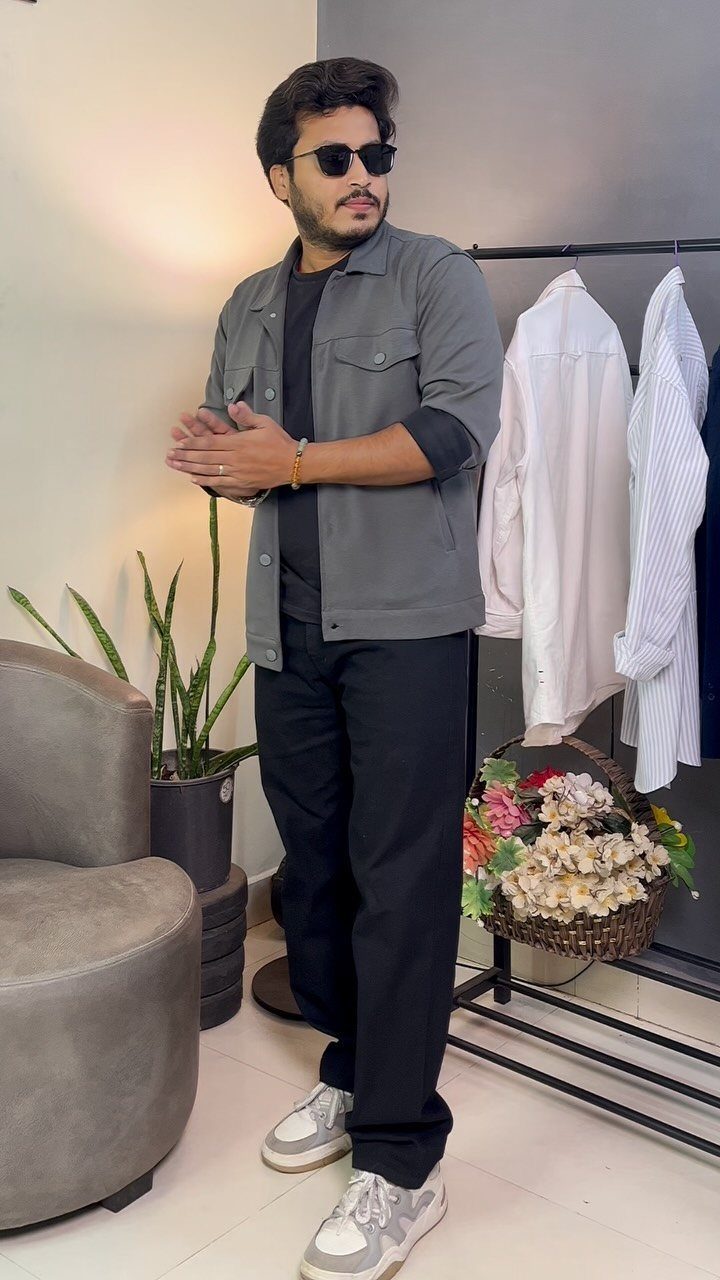 Man in a grey shirt, black t-shirt, black trousers, and sunglasses standing indoors.