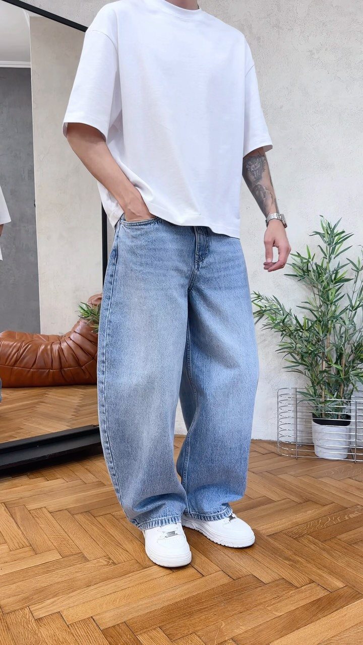 A man wearing an oversized white t-shirt, baggy light blue jeans, and white sneakers standing on a wooden floor.