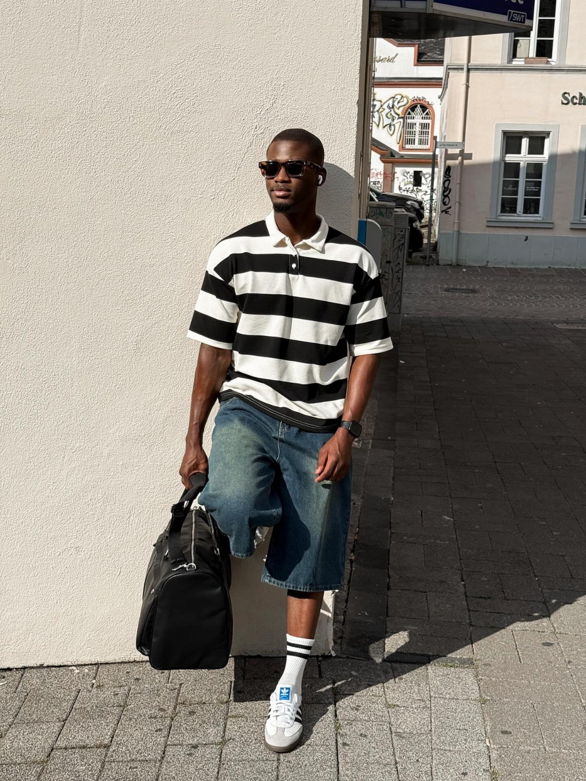 Man in a striped polo shirt and denim shorts standing against a white wall.