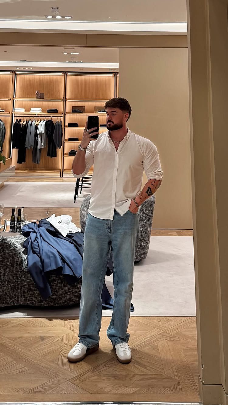 Man in a white shirt and light blue jeans posing for a mirror selfie.