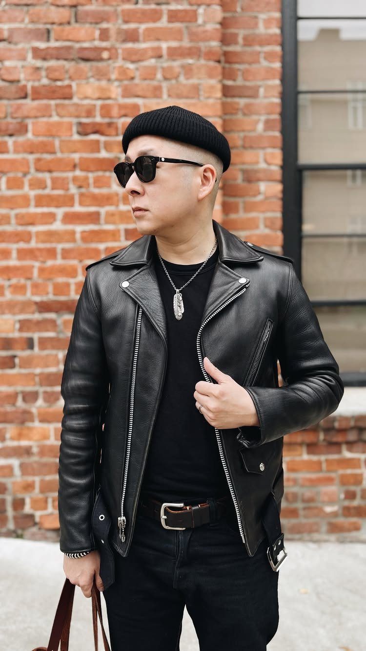 Man in a black leather biker jacket, black t-shirt, and sunglasses standing against a brick wall.