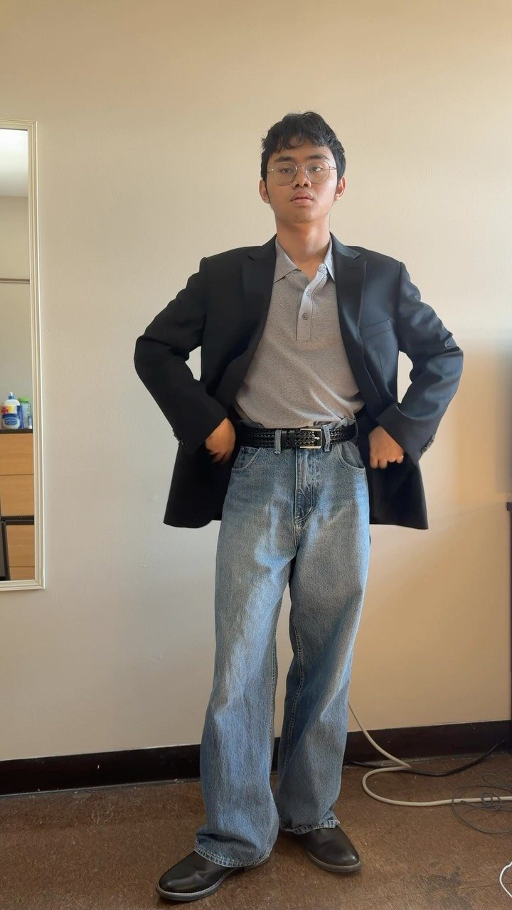 A man wearing a black blazer, grey polo shirt, and light blue jeans standing indoors.