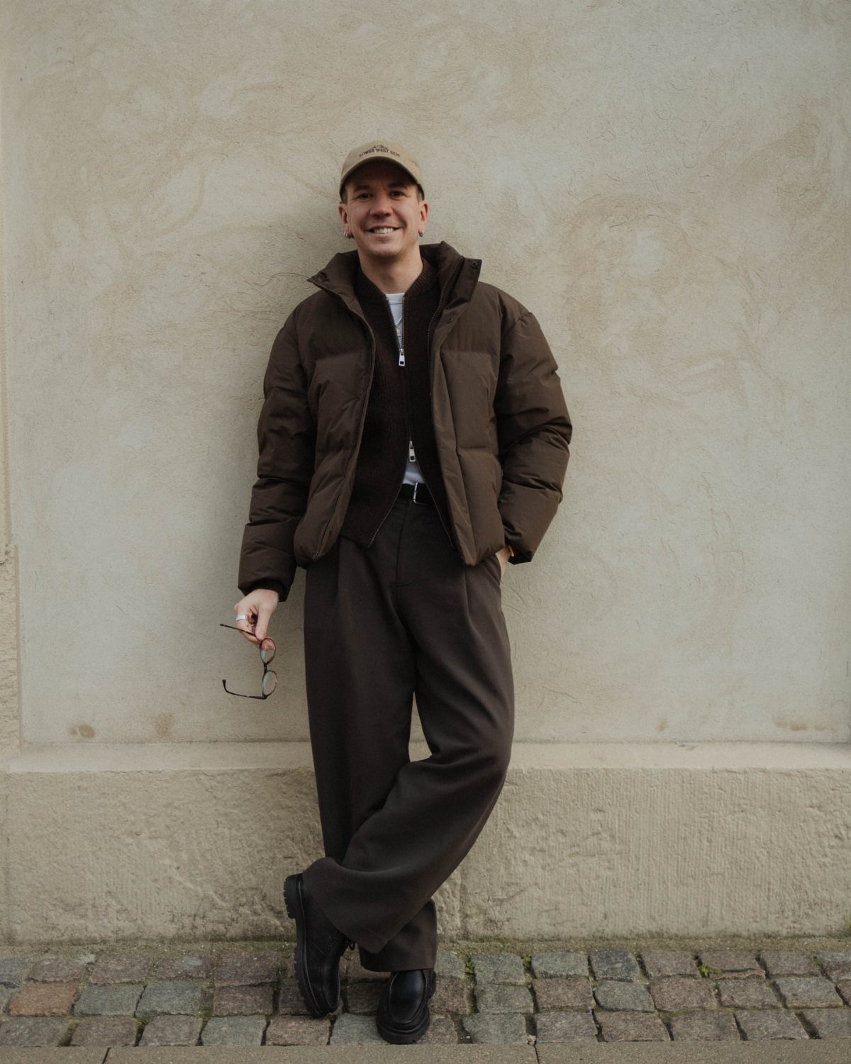 A man smiling while wearing a brown puffer jacket and dark brown trousers against a neutral wall.