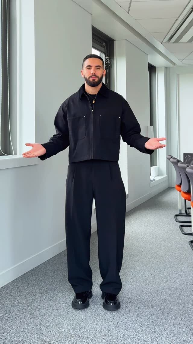 A man posing in a hallway wearing a black cropped zipper jacket and wide-leg black trousers.