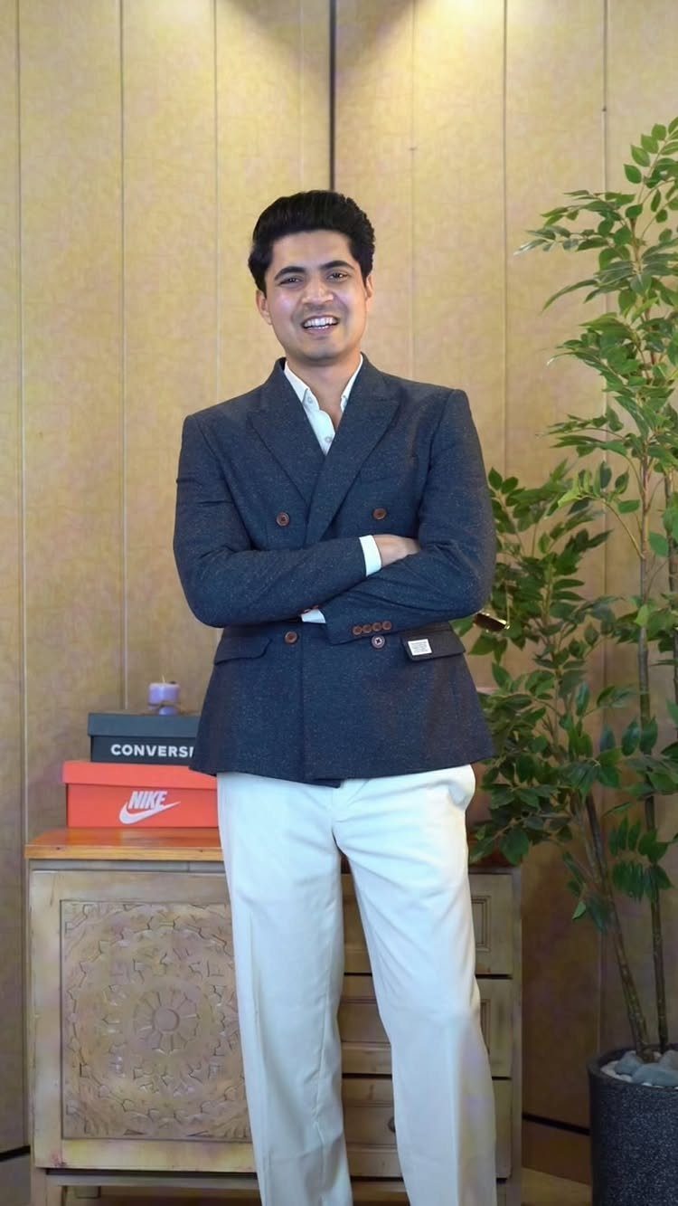 A man smiling in a dark grey double-breasted blazer and white trousers.
