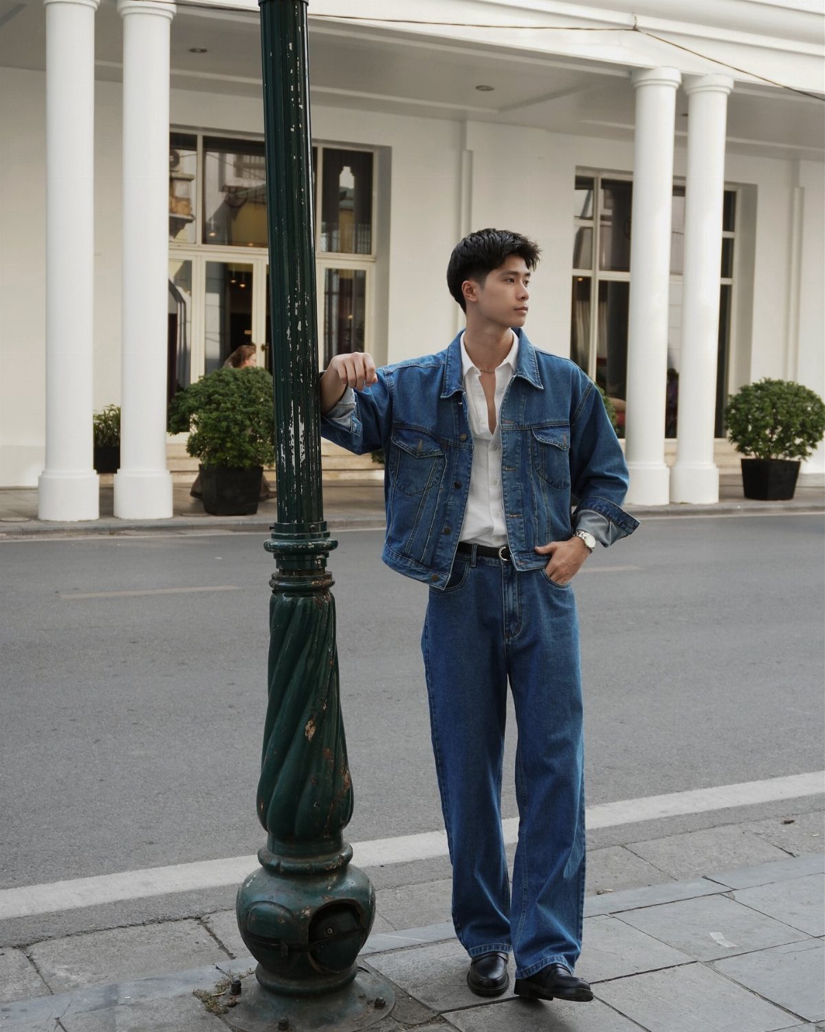 Man in a blue denim jacket and denim jeans leaning against a green lamp post.