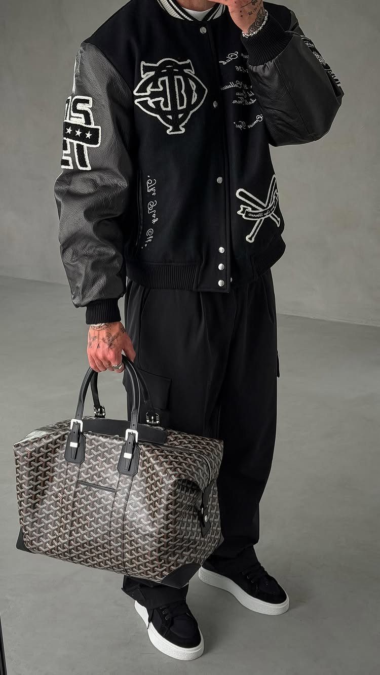 Man wearing a black and grey varsity jacket with black trousers and a patterned duffel bag.