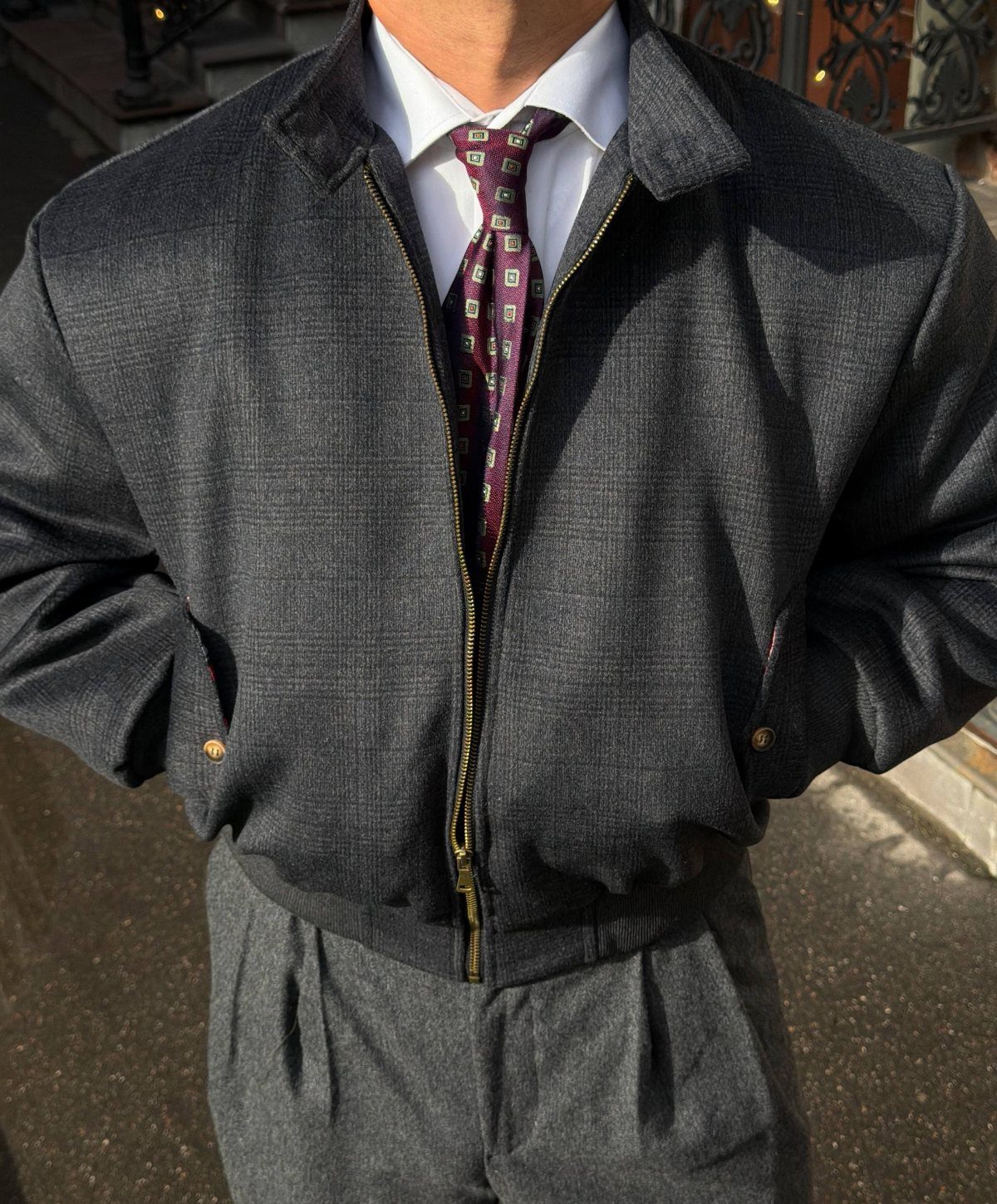 A man wearing a grey Harrington jacket, white shirt, and patterned purple tie.