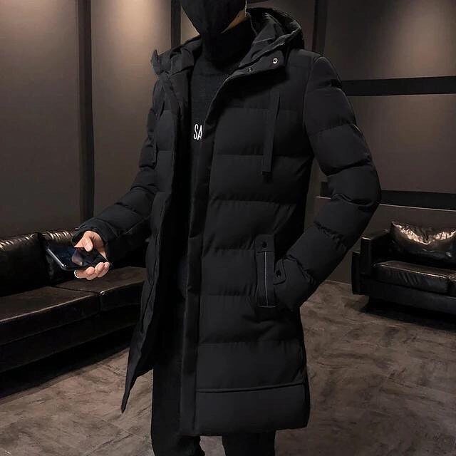 A man wearing a long black quilted puffer jacket with a hood standing in a dark room.