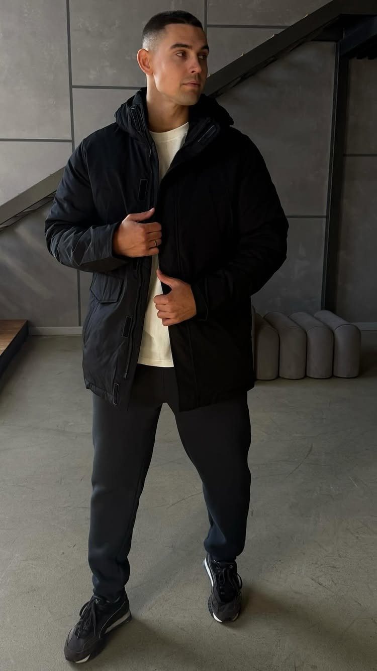 A man wearing a black hooded parka jacket, white t-shirt, and dark joggers standing indoors.