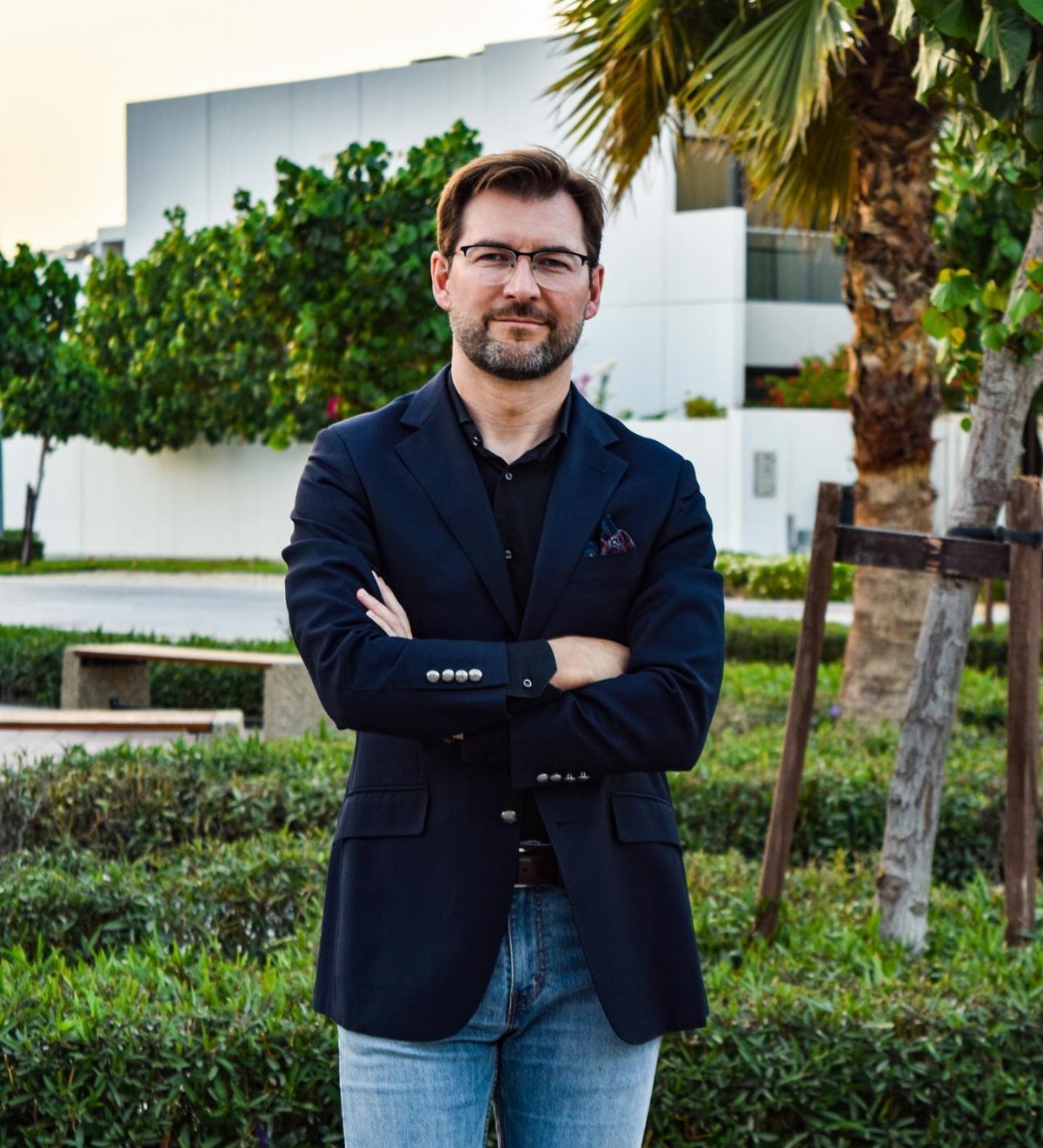 Man wearing a dark navy blazer and light blue jeans standing outdoors with arms crossed.