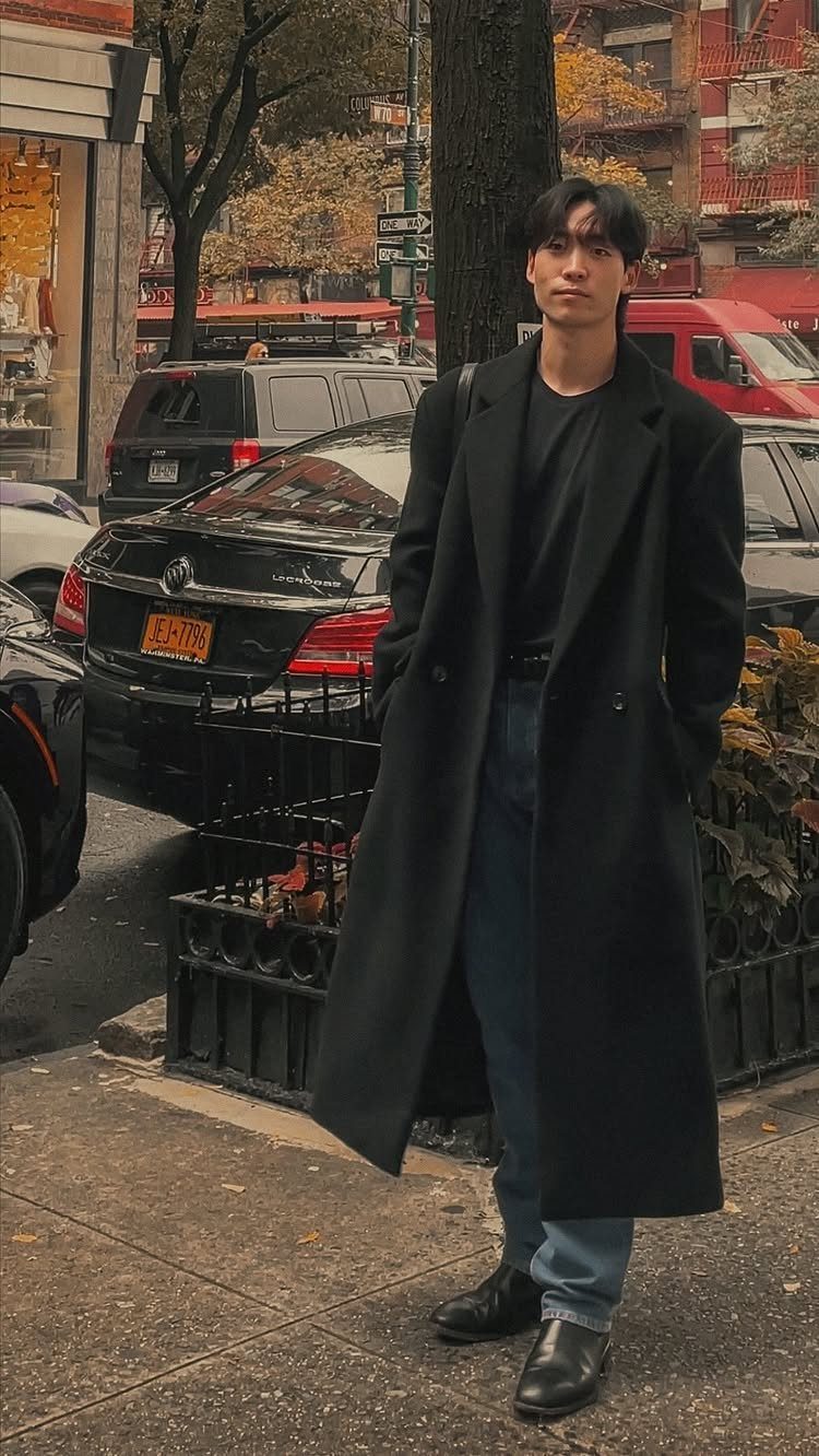 Man in a long black overcoat and blue jeans standing on a city street.