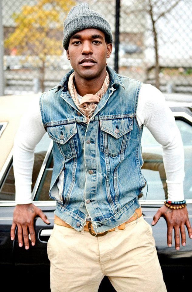 Man in a denim vest, white shirt, tan pants, and a patterned scarf leaning on a car.