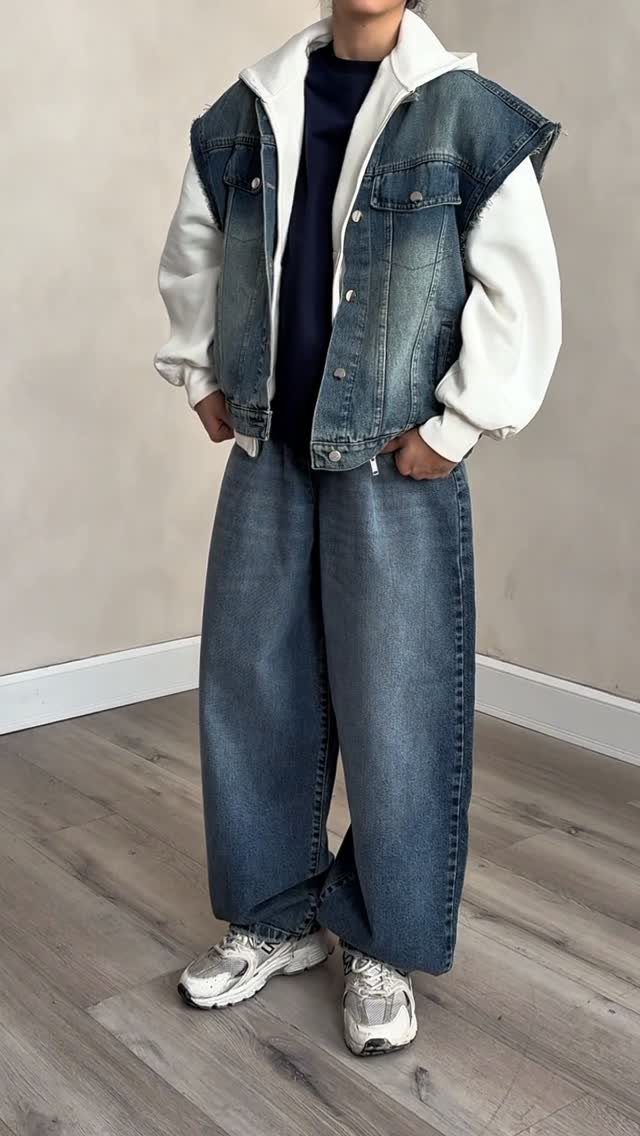 Person wearing a denim vest, white hoodie, wide-leg jeans, and grey sneakers.
