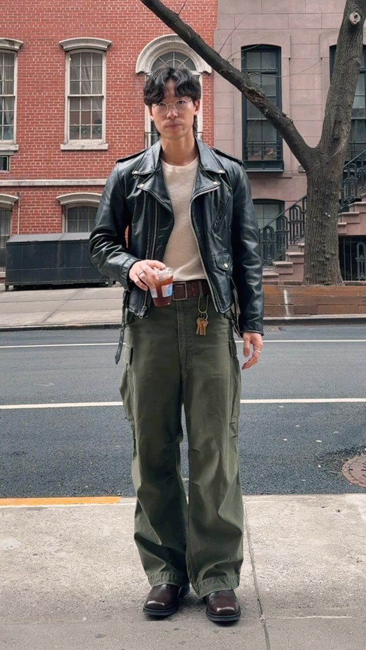 Man posing in a black leather jacket, cream shirt, and olive green cargo pants on a city street.