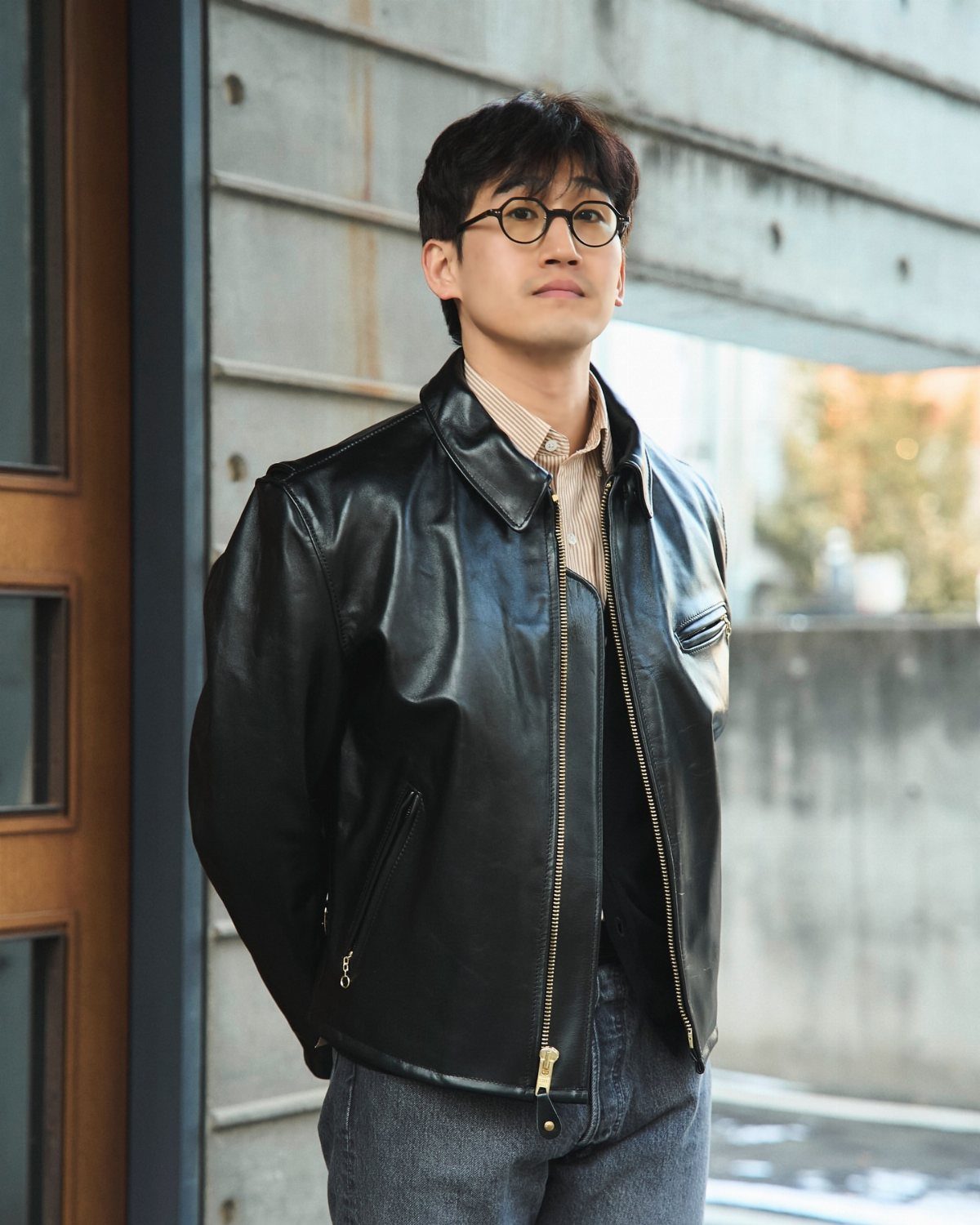A man wearing a black leather jacket and a striped shirt standing against a concrete wall.