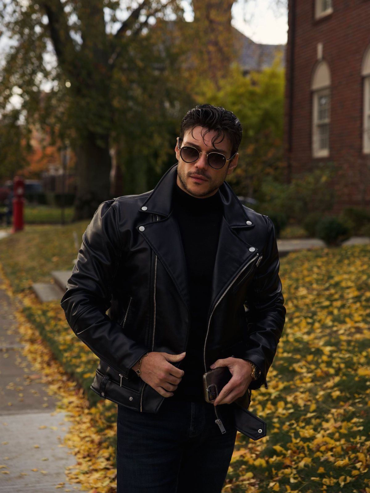 Man in a black leather jacket and sunglasses standing among autumn leaves.