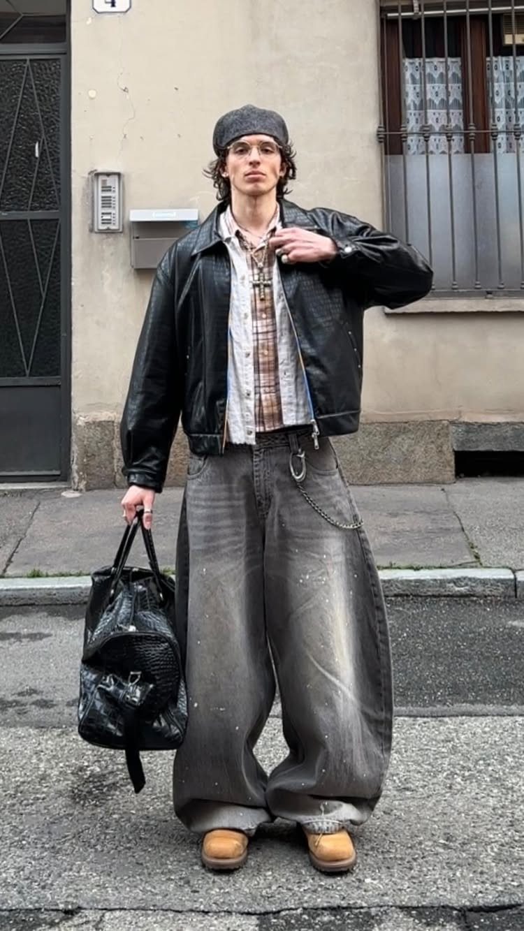 A man posing in a black leather jacket, plaid shirt, and wide-leg trousers.