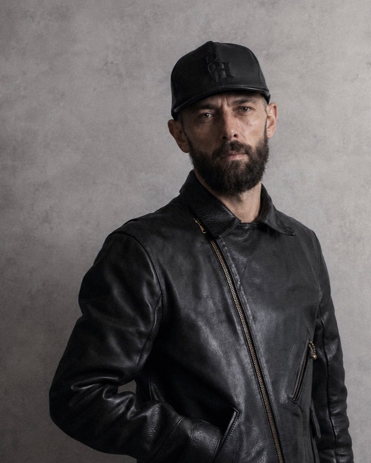 A bearded man wearing a black leather jacket and a matching black baseball cap.