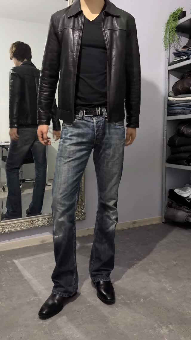 Man wearing a black leather jacket, black shirt, and blue jeans standing in a room.