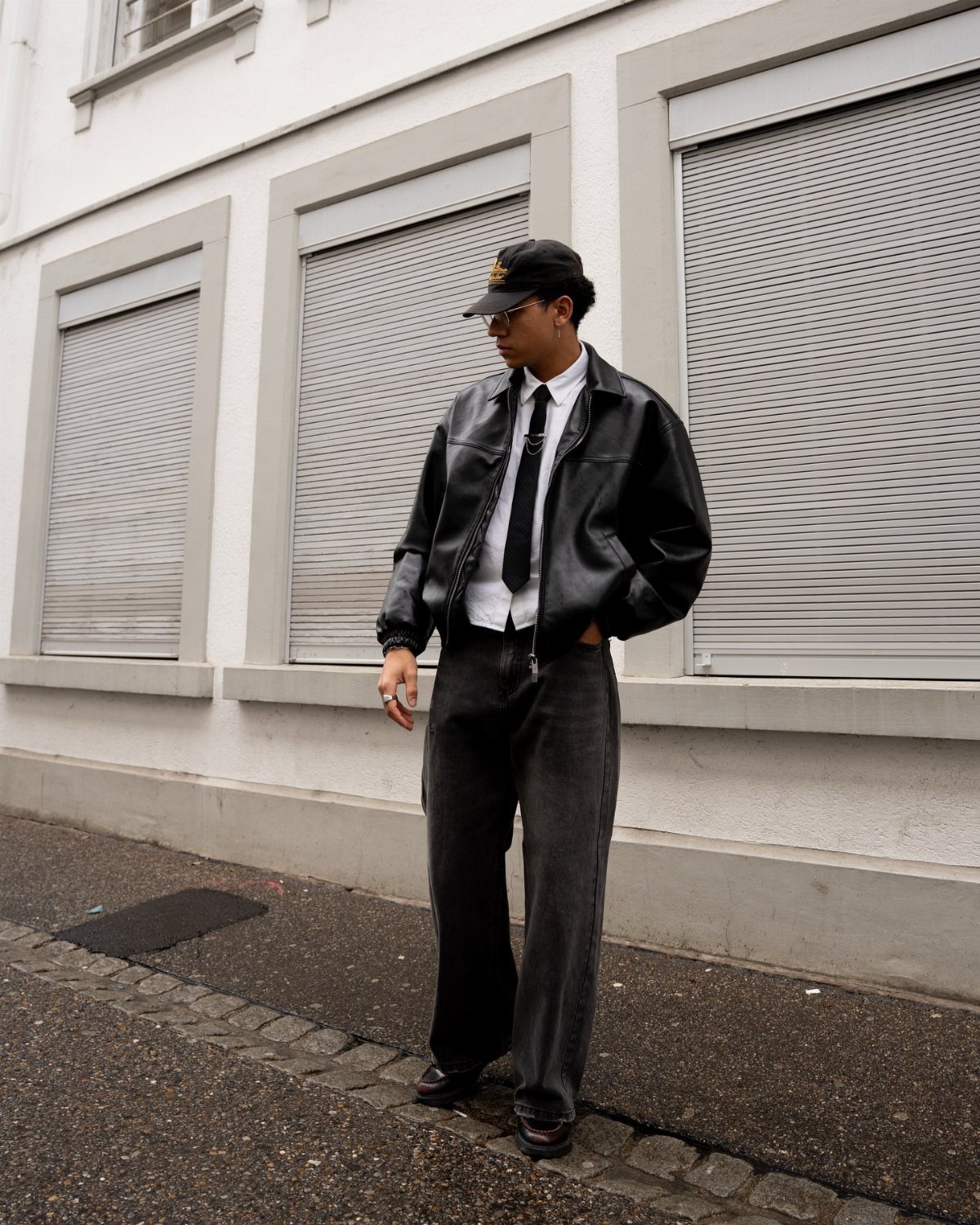 A person in a black leather jacket, white button-up shirt, and black tie standing outdoors.