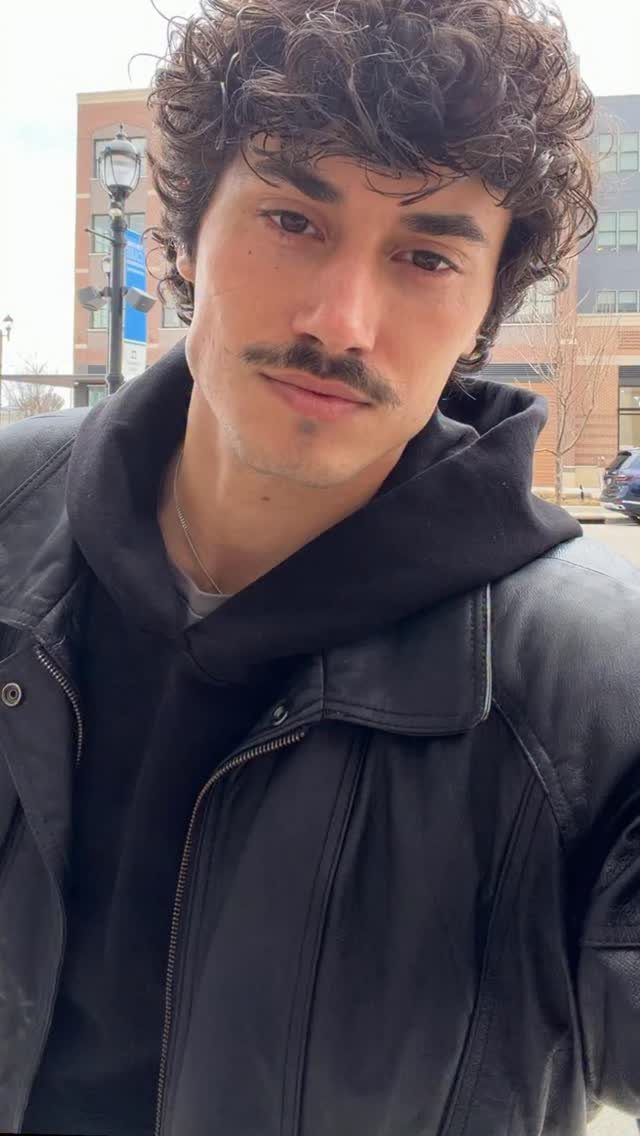 A young man with curly hair wearing a black leather jacket over a black hoodie.