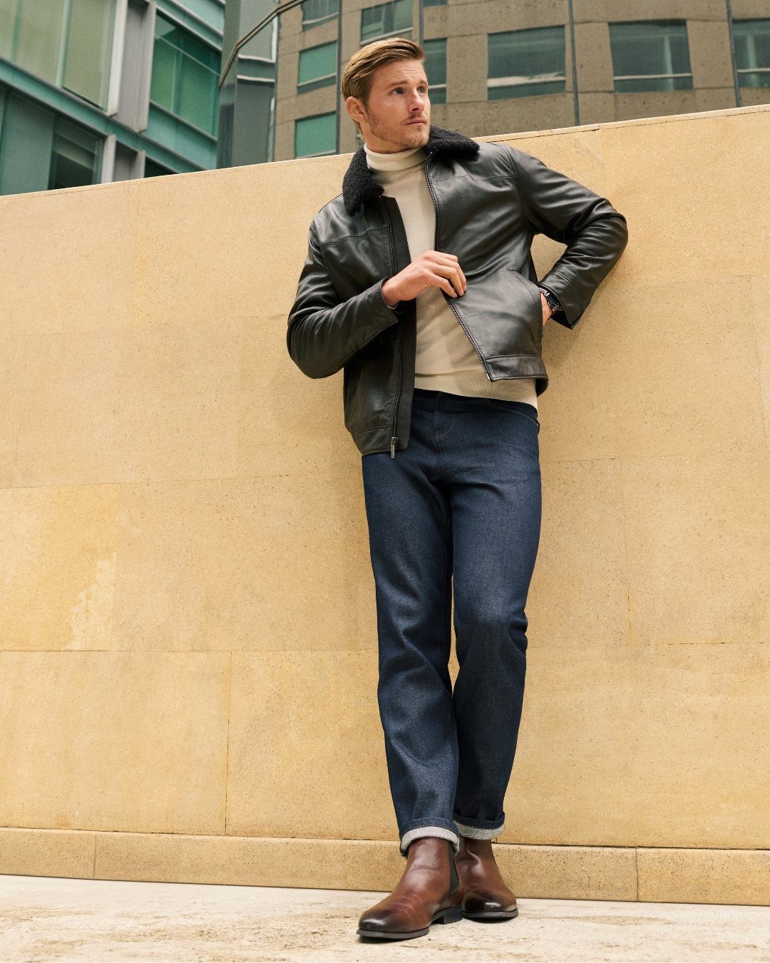 Man wearing a black leather jacket over a cream turtleneck sweater and dark blue jeans.