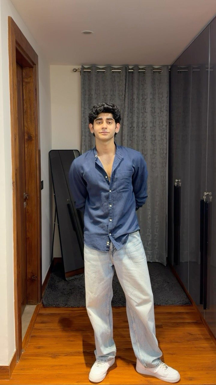 A young man posing in a blue shirt, light blue jeans, and white sneakers.