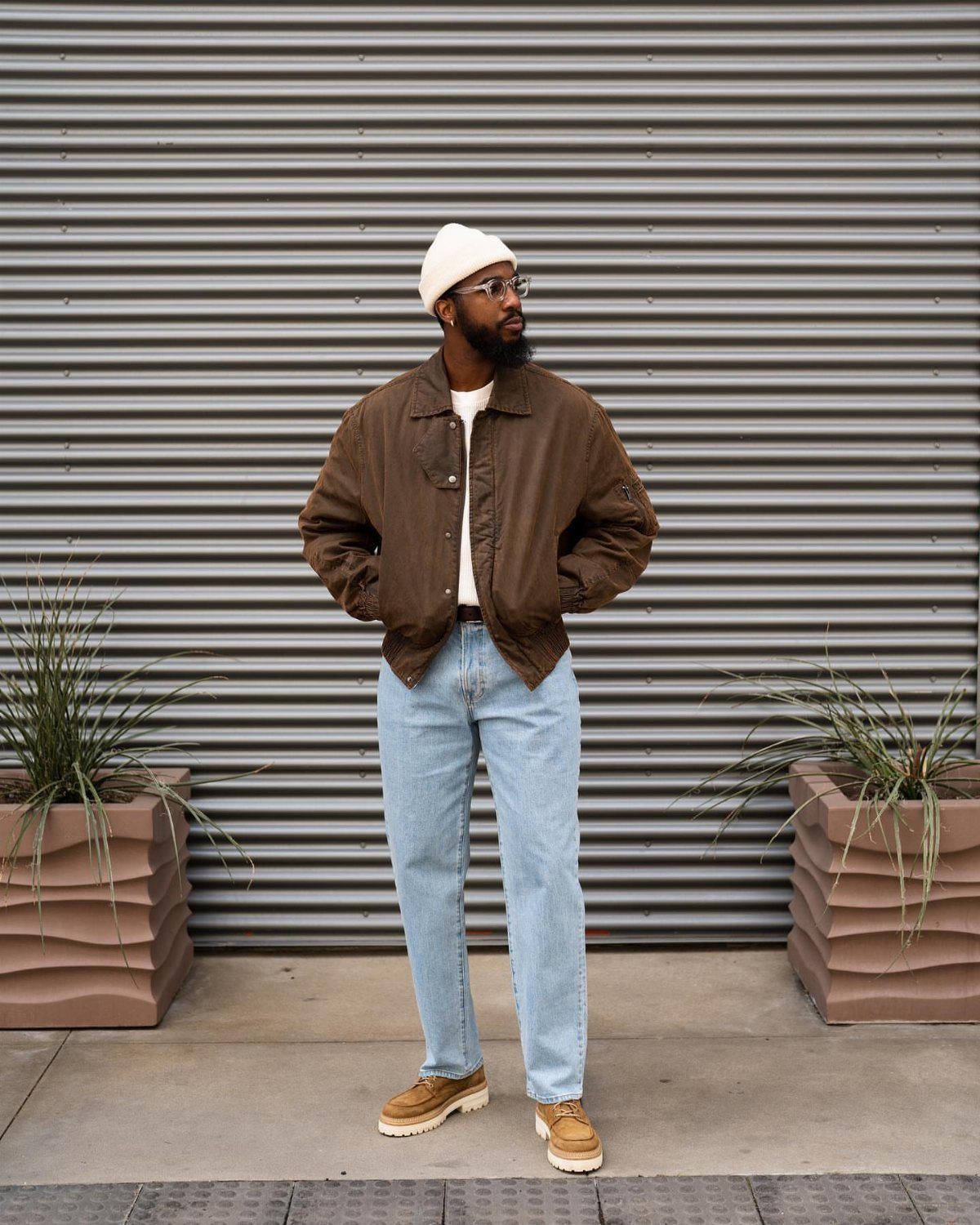 Man in a brown bomber jacket and light blue jeans standing against a corrugated wall.