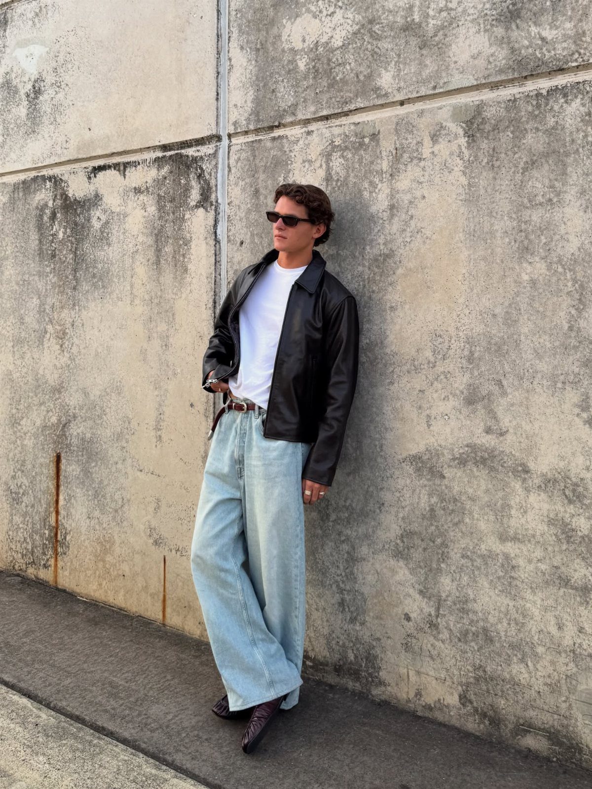 Man in a black leather jacket, white t-shirt, and light blue jeans leaning against a wall.