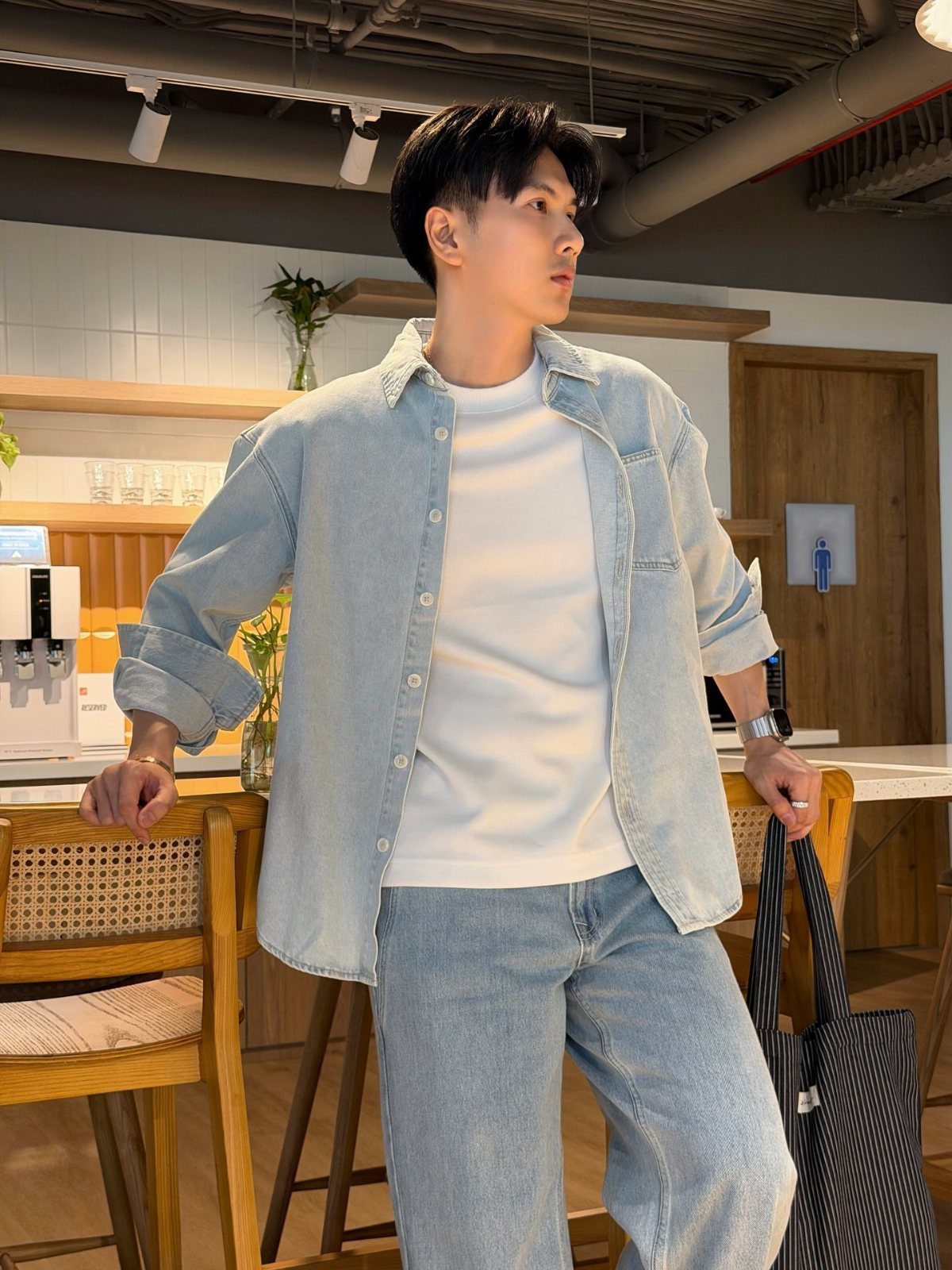 Man in a white t-shirt, light blue denim shirt, and light blue jeans standing indoors.