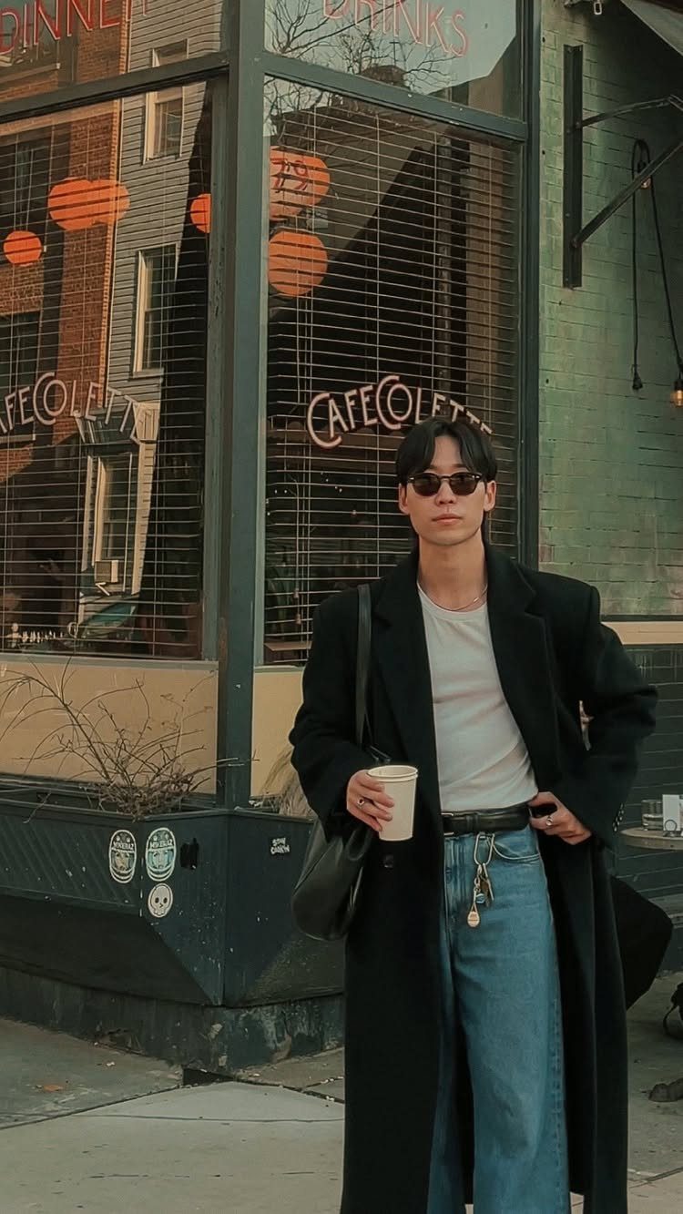 Man in sunglasses, white t-shirt, black coat, and light blue jeans standing outdoors.