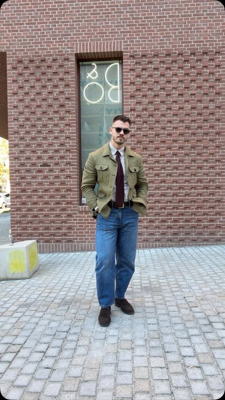 Man in olive jacket, white shirt, tie, and blue jeans standing on cobblestones.