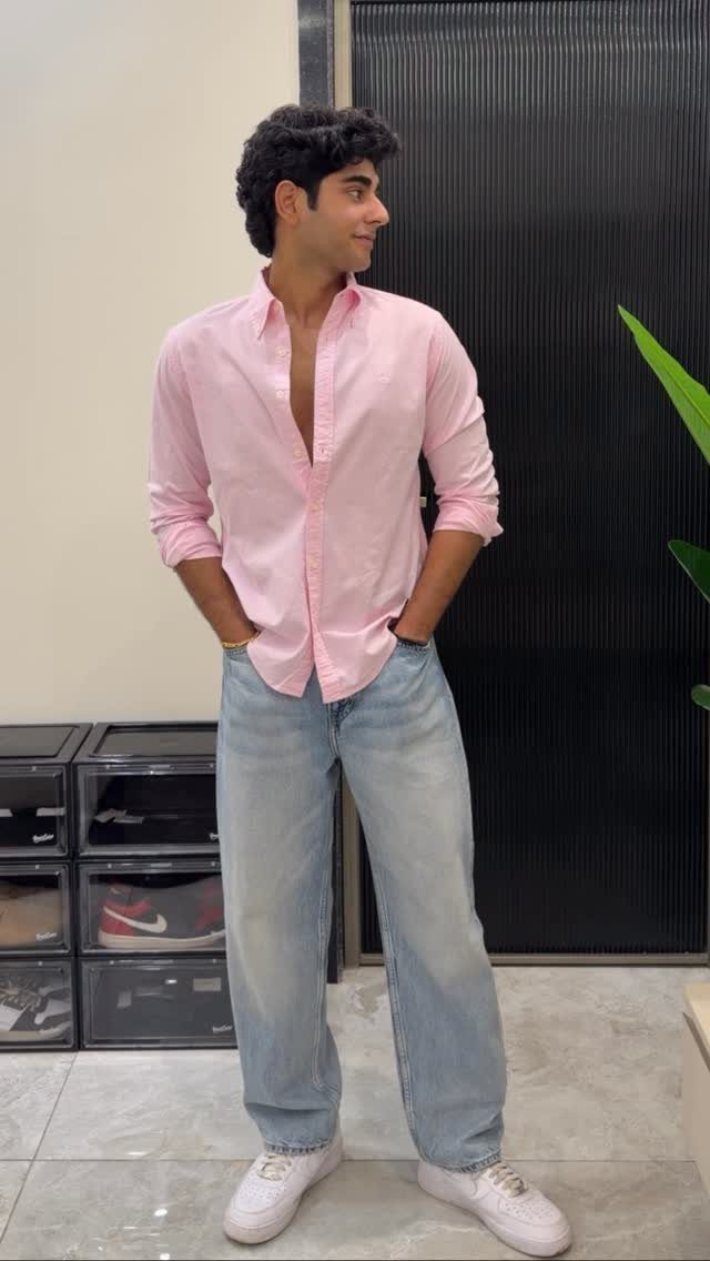 A man wearing a pink shirt and light blue jeans standing indoors.