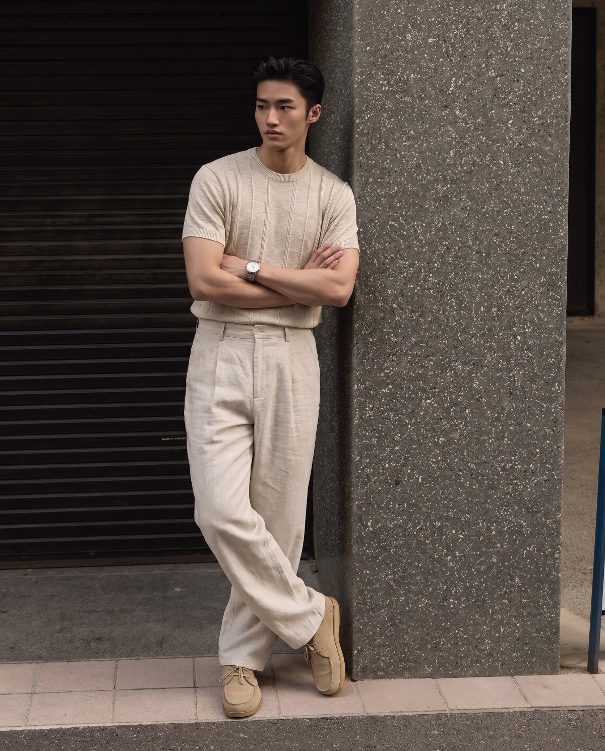 Man in a cream knit sweater and linen pants leaning against a stone pillar.