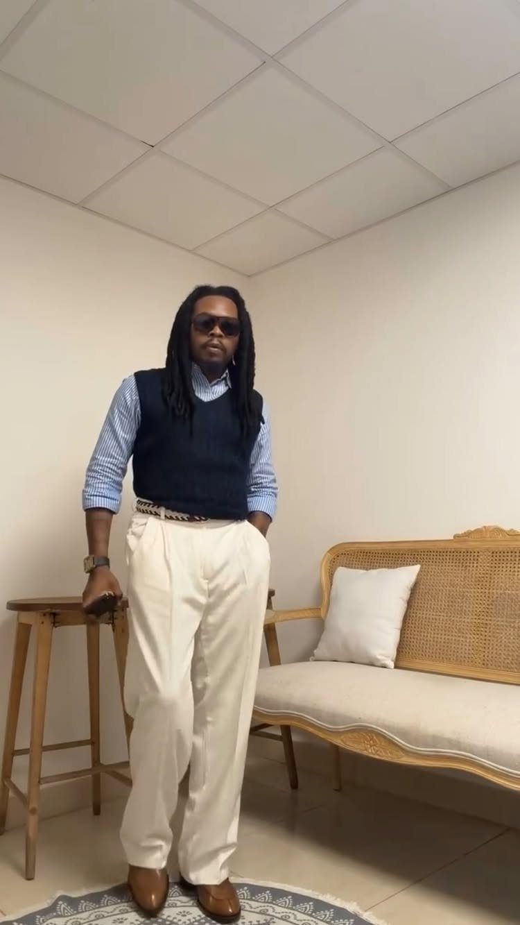 Man in a blue button-up shirt, navy sweater vest, and white linen pants standing indoors.