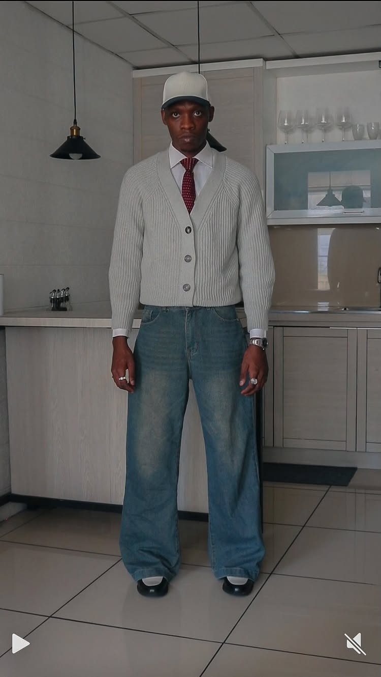 Man in a white cap, grey cardigan, red tie, and wide blue jeans standing in a kitchen.